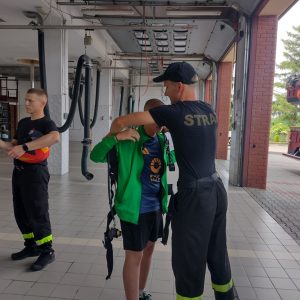 Dwóch strażaków pomaga uczniom przymierzyć butlę z tlenem.