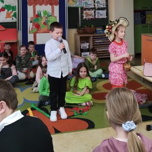 Dzieci siedzą na dywanie. Jeden chłopiec trzyma mikrofon, dziewczynka ubrana jak pani wiosna.