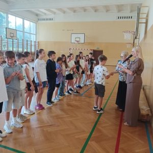 Pani dyrektor wręcza chłopcu kartę rowerową, zadowolone dzieci stoją na żółtej linii.
