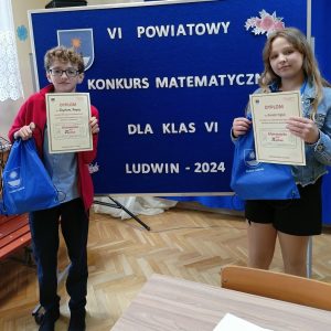 reprezentaci naszej szkoły Krystian i Amelia stoją przy tablicy z tytułem konkursu. W ręce trzymają dyplomy oraz niebieskie worki z nagrodami