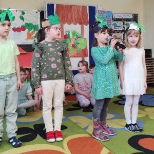 Dzieci stoją w rzędzie, dziewczynka trzyma mikrofon, każde z dzieci recytuje swoją rolę inscenizując wiersz „Pomidor".
