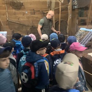 Dzieci stoją przed przewodnikiem a w tle eksponaty muzealne